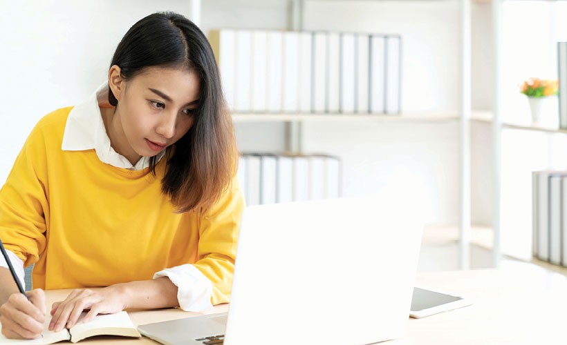 A female student writes on a notepad, sitting with her laptop in a bright room